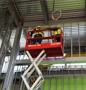 Two workers in safety vests and hard hats stand on a red MEC Micro 26 scissor lift, working on a metal ceiling frame inside an industrial site .