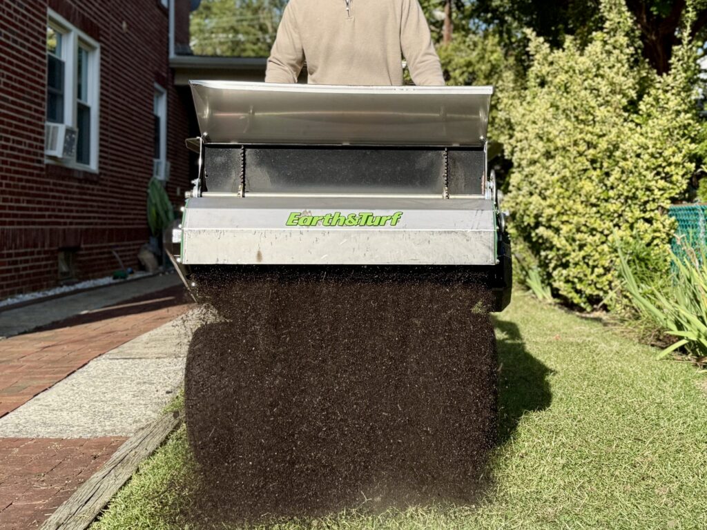 Person standing behind a metal Earth & Turf soil spreader dumping a large pile of dark compost onto a driveway beside a brick house and green lawn.