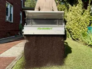 Person standing behind a metal Earth & Turf soil spreader dumping a large pile of dark compost onto a driveway beside a brick house and green lawn.
