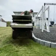 Lawn care worker in a green shirt dumps material from a cart onto a lawn beside a gravel play area with a swing set nearby.