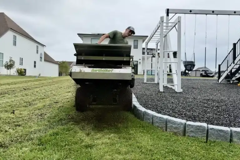 Lawn care worker in a green shirt dumps material from a cart onto a lawn beside a gravel play area with a swing set nearby.