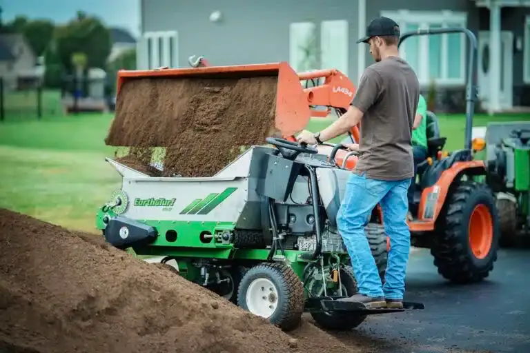 Man guiding soil into a green-orange soil screener attached to a tractor on a residential street.