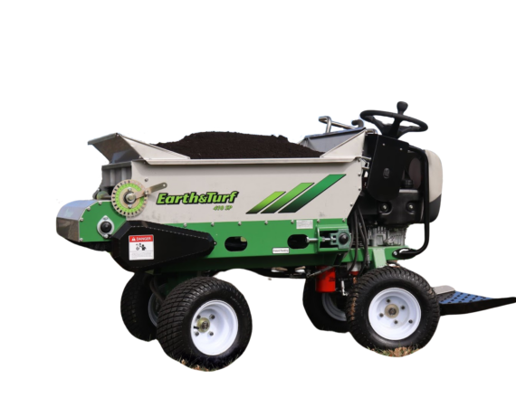 Ride-on turf aerator labeled 'Earth & Turf' with a dirt-filled hopper on top, parked on grass beside a tree.