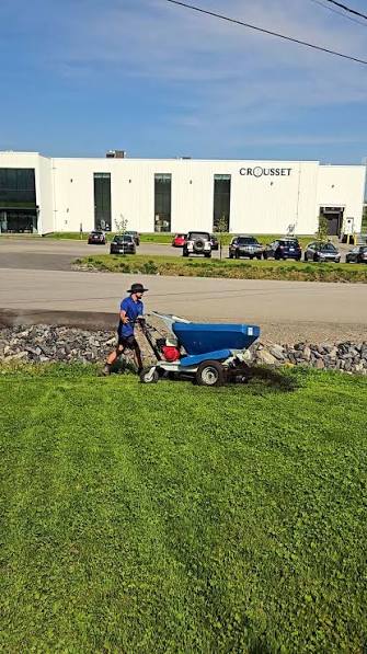 Person in a blue shirt and hat pushes a blue lawn spreader on a grassy area near a road, with a white commercial building and parked cars in the background.